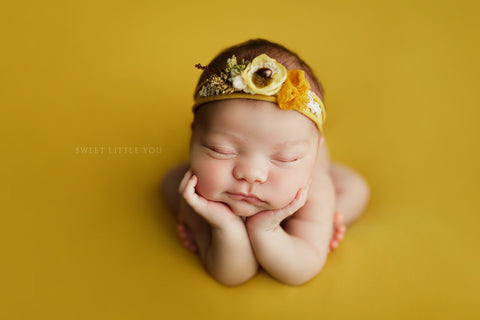 Newborn baby professional photography photo. Baby is in "froggy pose." She is styled on a mustard DreamSoft backdrop and wearing a coordinating photography prop headband.