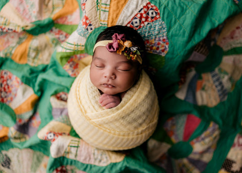 Image of Black newborn baby from a professional photography session.  Baby is laying on an antique quilt piece. Baby is swaddled in a "Soleil" photo prop wrap with a coordinating headband.