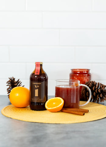 A photo of a brown glass bottle of Curious Number 1 beside a glass mug full of a deep red drink. These are surrounded by oranges, pinecones, and a seasonal candle.