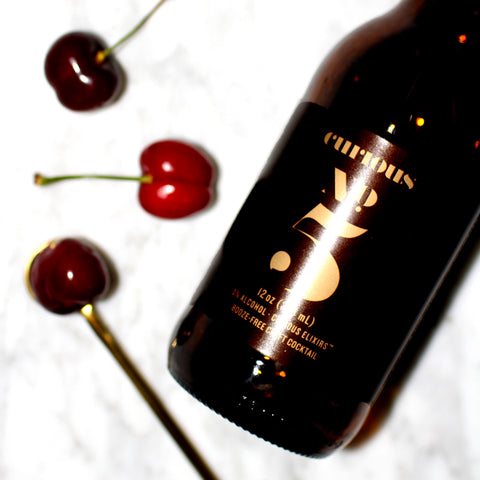 A photo of a brown glass bottle of Curious Elixir Number 5 laying beside three cherries on a white marble surface. 