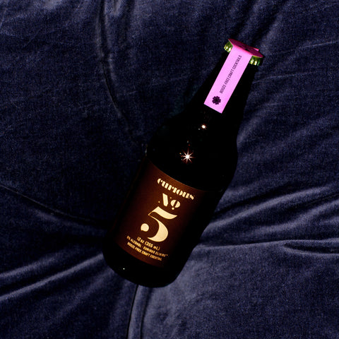 A photo of a brown glass bottle of Curious Elixir Number 5 laying on a dark blue velvet cushion.