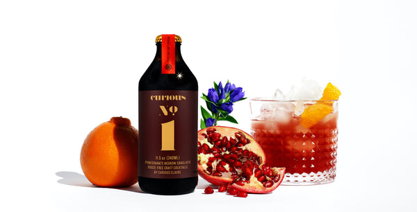 A photo of a brown glass bottle of Curious Number 1 beside a glass containing the same drink. These are surrounded by an orange, half a pomegranate, and a plant with purple flowers.