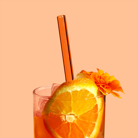 A photo of a glass with an orange beverage in it. The drink is garnished with a round slice of orange and an orange flower. An orange straw sticks out of it. The background is a pale peach color. 