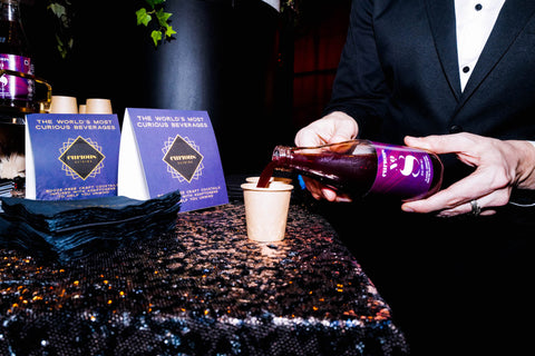 A photo of a bottle of Curious Number 8 being poured into small paper cups. The beverage is a rich purple color, and pamphlets about Curious Elixirs, also purple, are on the table in the background.