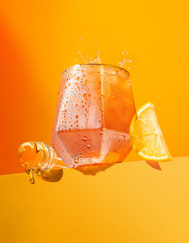 A photo of a glass of Curious. The drink is a light orange color, and a lemon slice and honey stick lean against the side of the glass. The background is orange and yellow.