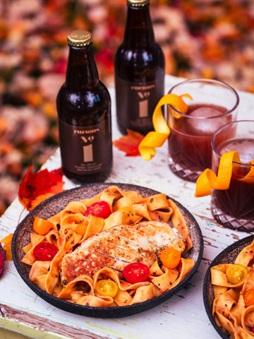A photo of two brown glass bottles of Curious Number 1 beside a dish of a creamy tomato pasta dish with grilled chicken and cherry tomatoes on top. In the background, a landscape of fallen autumn leaves is visible.