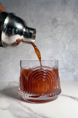 A silver drink shaker pouring a reddish-brown drink into a short glass full of ice.