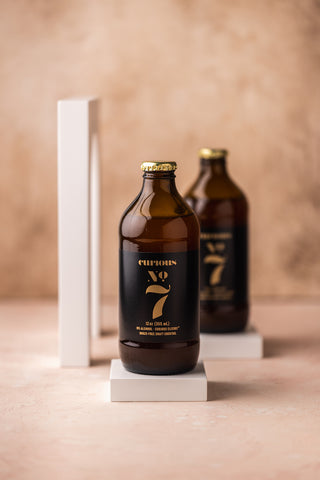 A photo of two brown glass bottles of Curious Number 7 sitting on small white blocks.