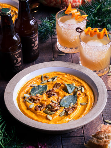 A photo of a bowl of pumpkin soup sitting in front of two bottles of Curious Number 2, as well as two glasses containing the same drink, garnished with orange peels.