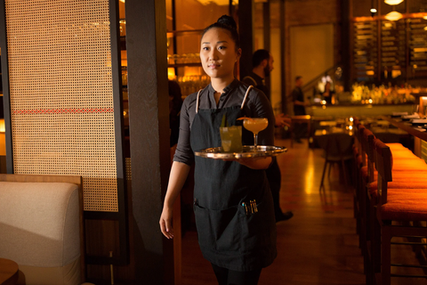 Female cocktail server in a charcoal shirt and black canvas apron made by Stock