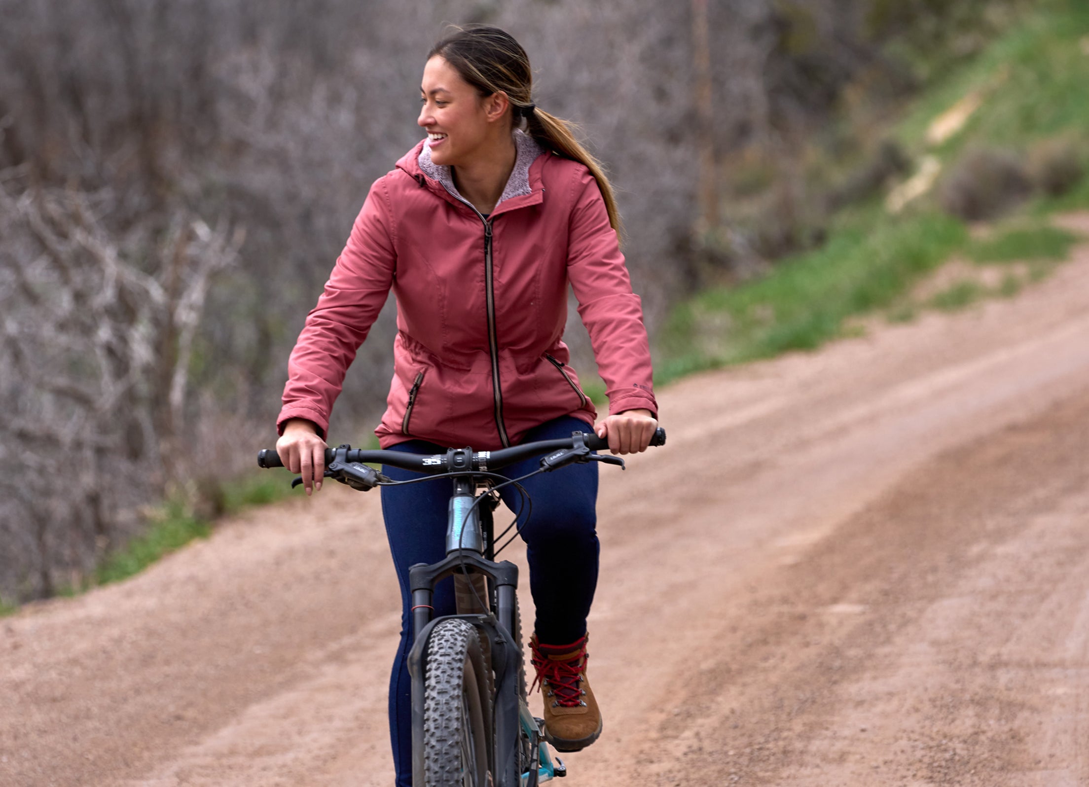 A woman rides a bike down a mountain trail wearing a pink Free Country Windshear jacket.