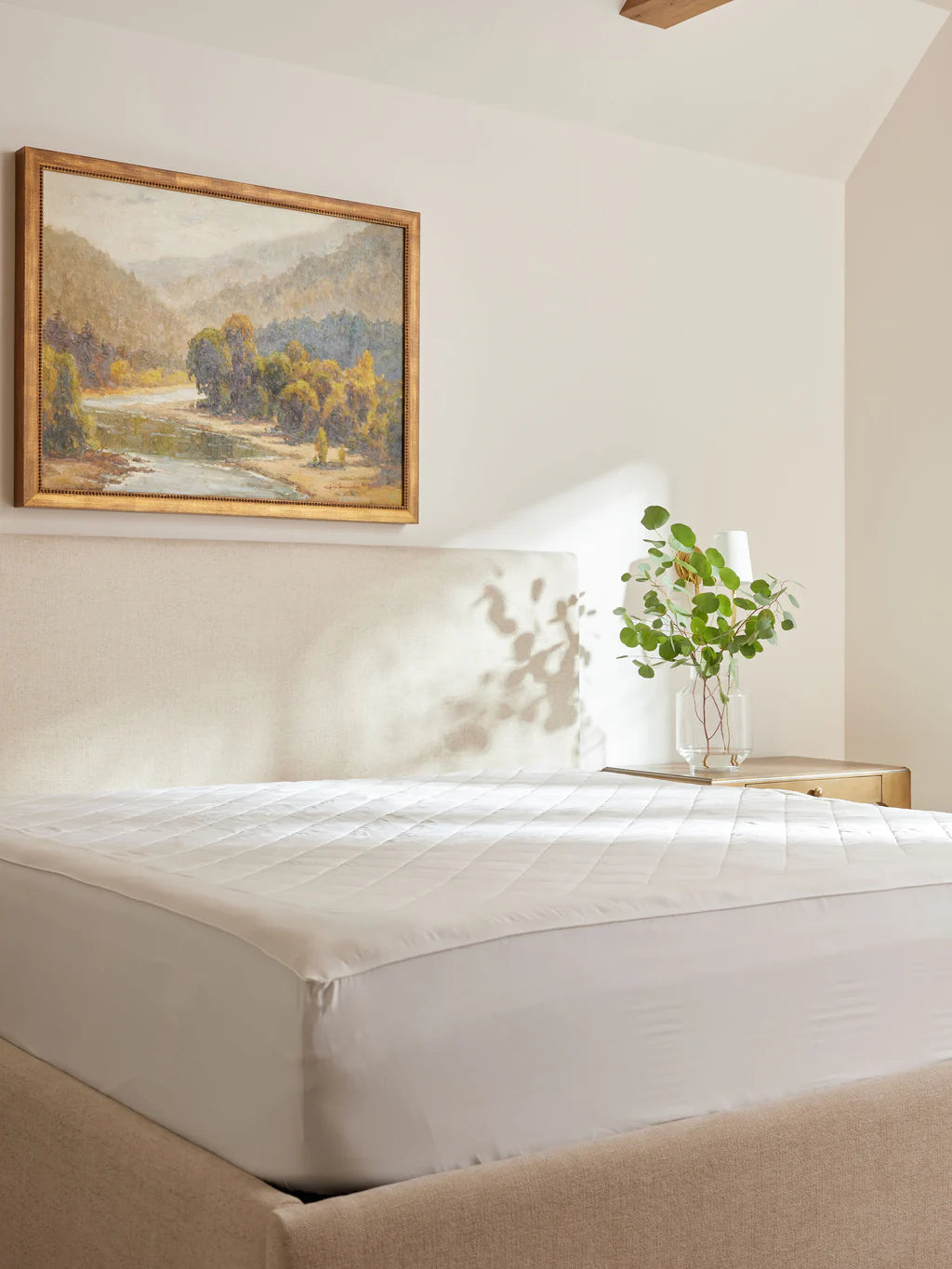 Bedroom with a neatly made bed, wall art, and a vase with leaves on a bedside table.