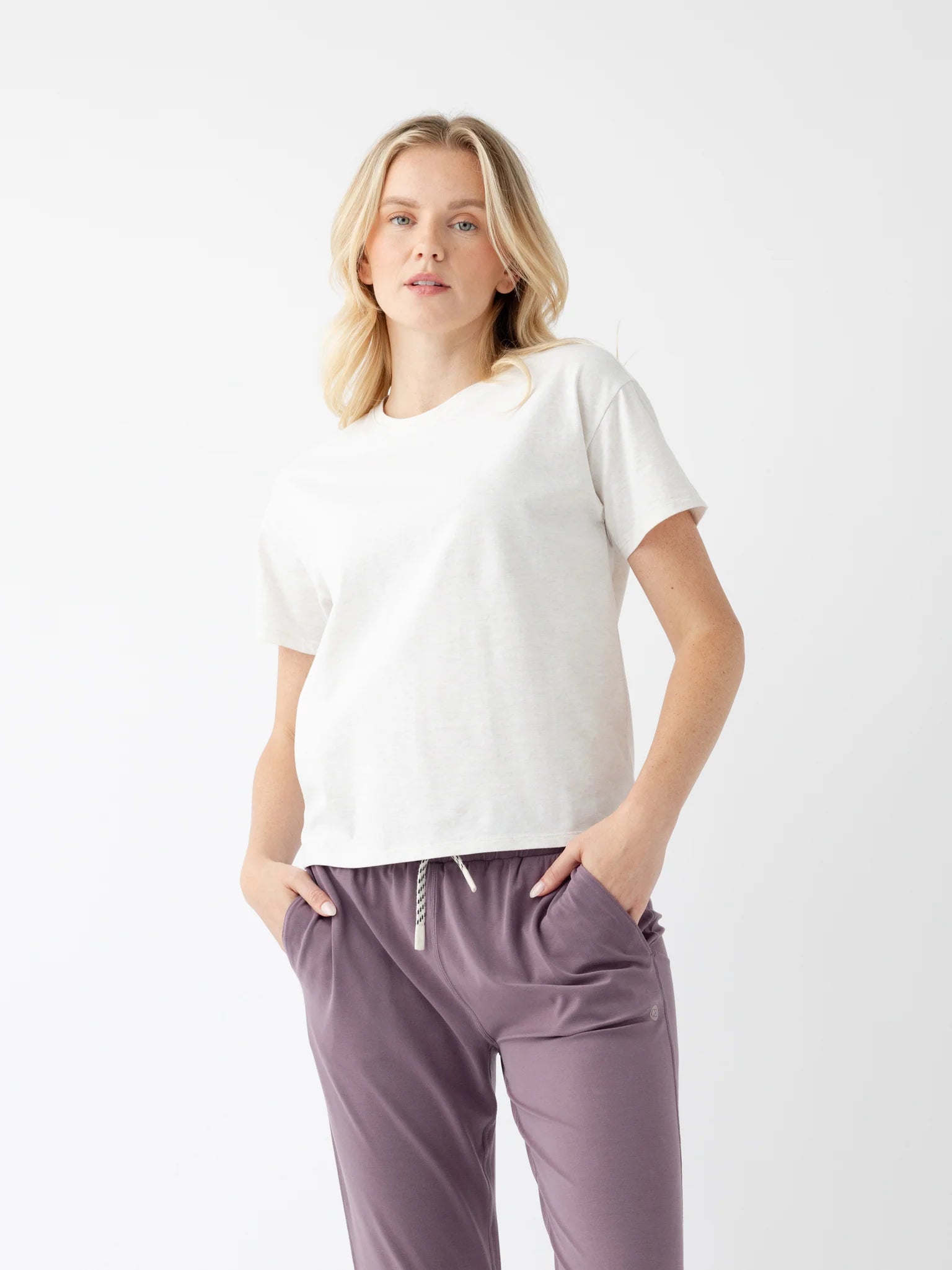 Woman wearing white t-shirt and purple pants with hands in pockets against plain background.