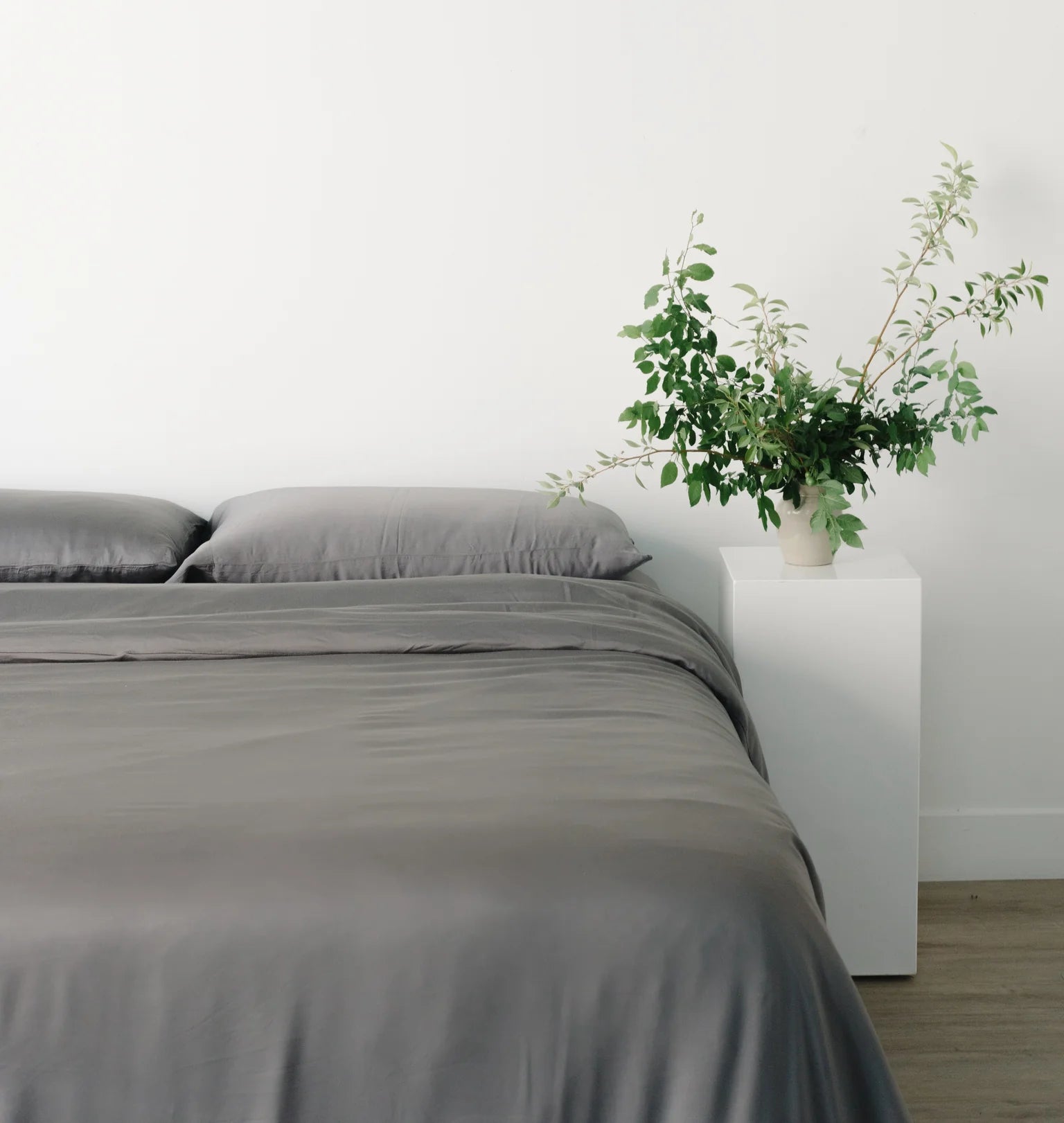 Minimalistic bedroom with grey bedding and a potted plant on a white nightstand.