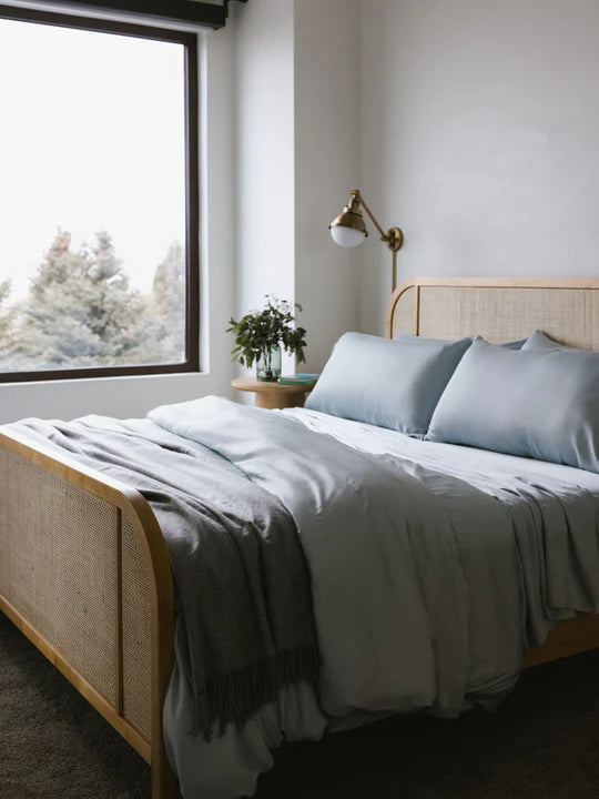 A cozy bedroom with a made bed, large window, and a small plant on the bedside table.