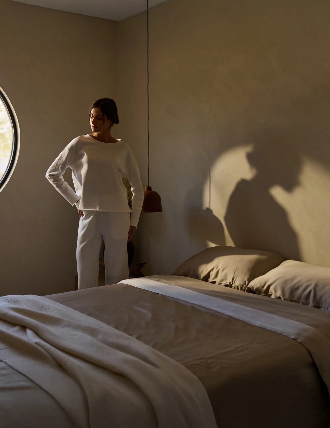 Person standing in a softly lit bedroom with a round window, casting a shadow on the wall.