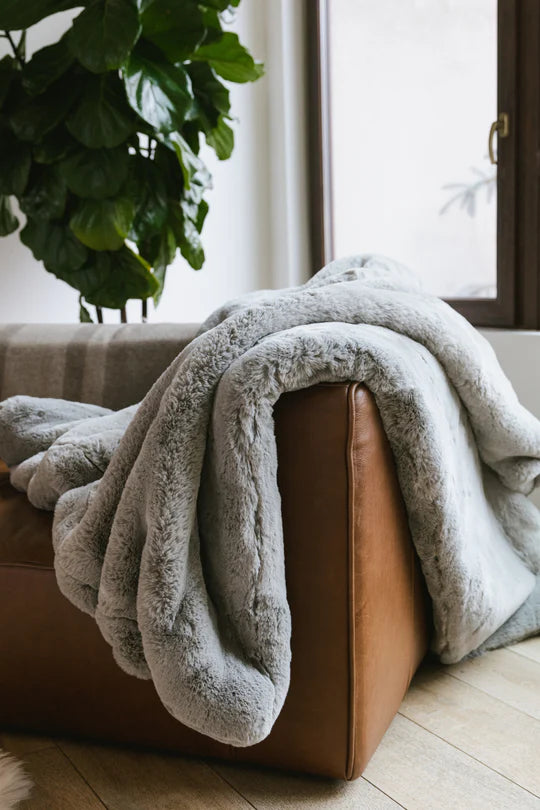 A plush grey blanket draped over a brown leather couch near a window and a green plant.