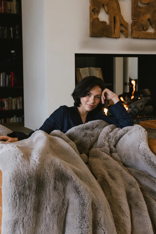 Person relaxing on a couch with a large, fluffy blanket near a bookshelf and fireplace.