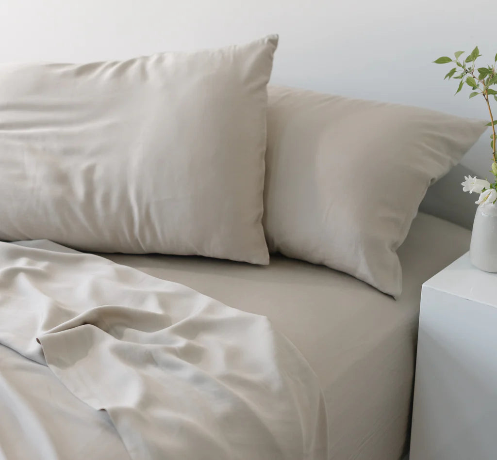 Bed with light gray sheets, pillows, and a small plant on a white nightstand.