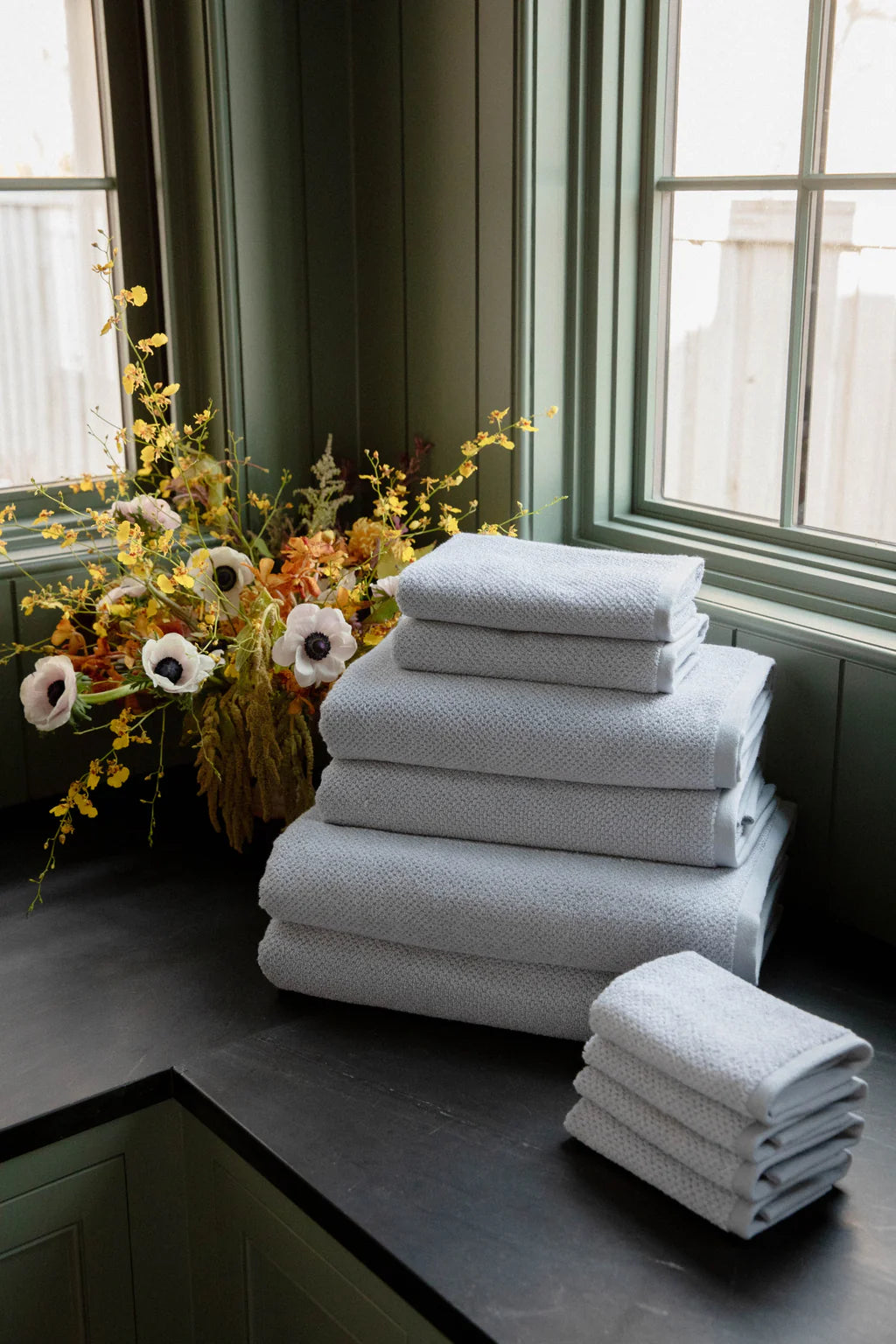 A stack of white towels next to a floral arrangement on a dark countertop by a window.