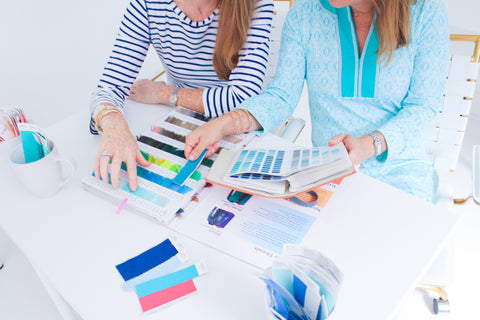 Women in blue dresses looking at colorful swatches & fabrics on white desk