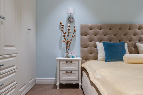  A bed with a leather headboard and upholstered frame, decorated with pillows and a comforter, next to a white bedside table with plants and other decorations.