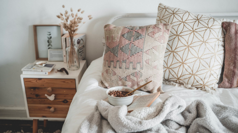 A bowl of cereal and a book on top of a mattress, decorated by pillows, with a metal bedframe in view next to a bedside table filled with ornaments