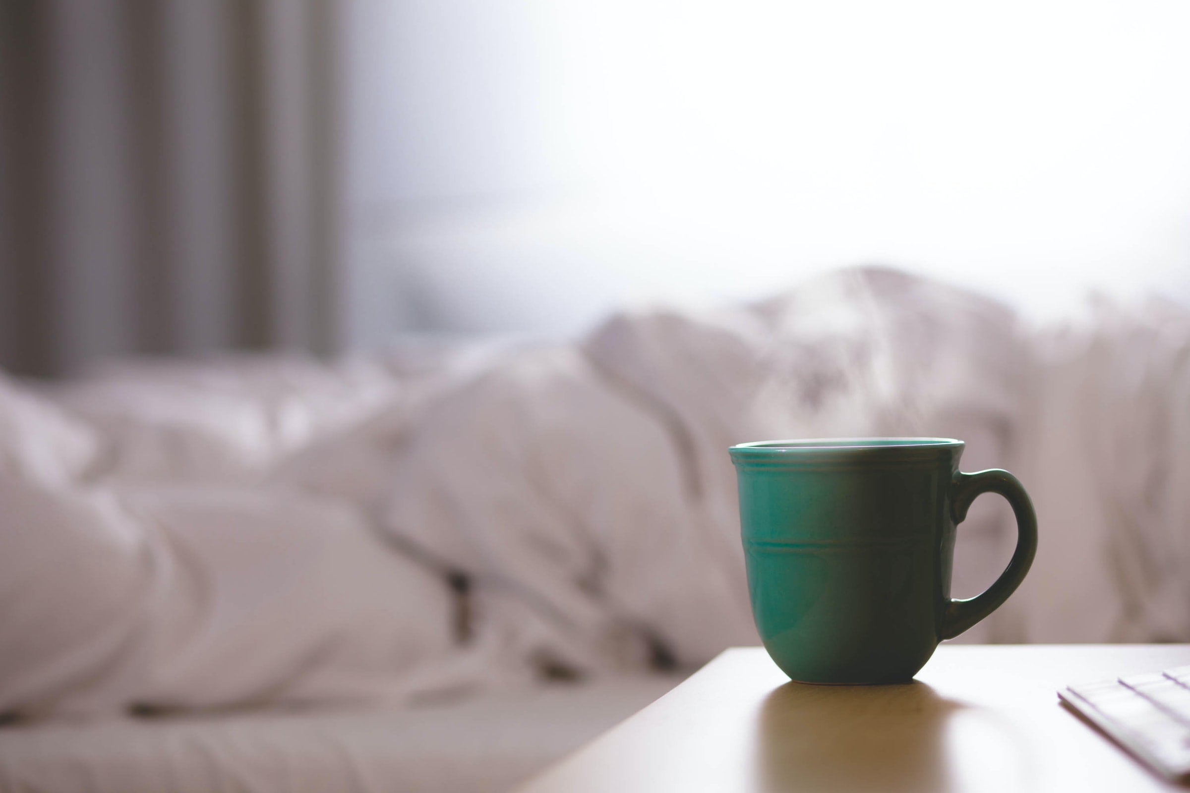 A green mug with steam coming out of it atop a bedside table, with the view of an unmade bed in the background