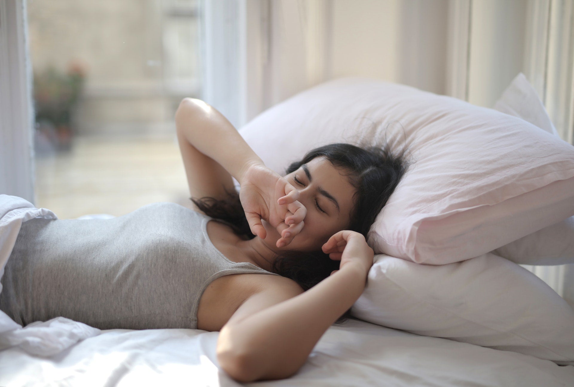 A girl lying in her bed, next to her window, with her palm covering her mouth as she yawns