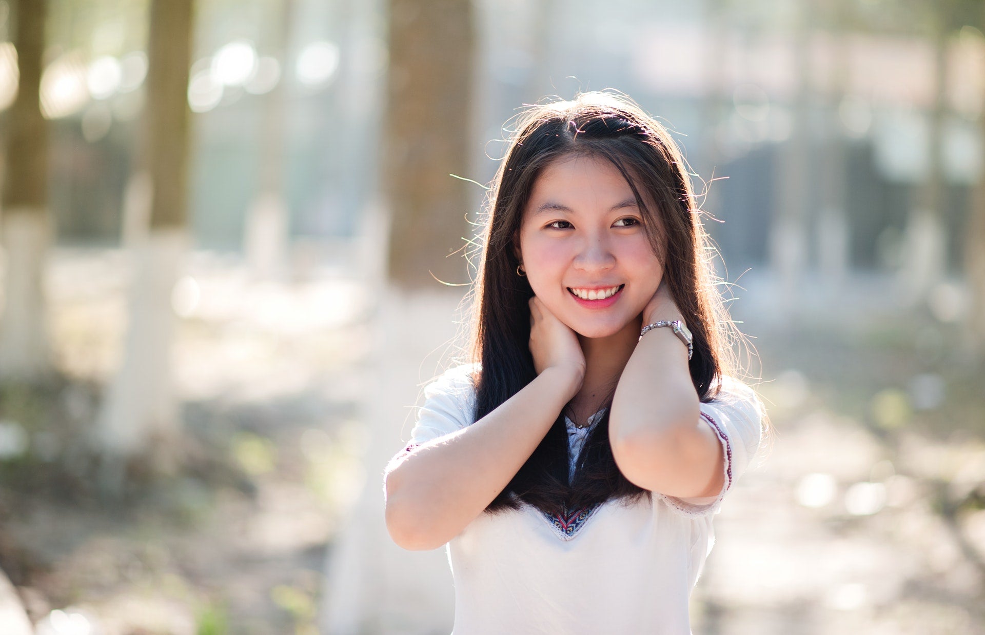 A girl dressed in white outdoors, smiling while reaching her hands to the back of her neck