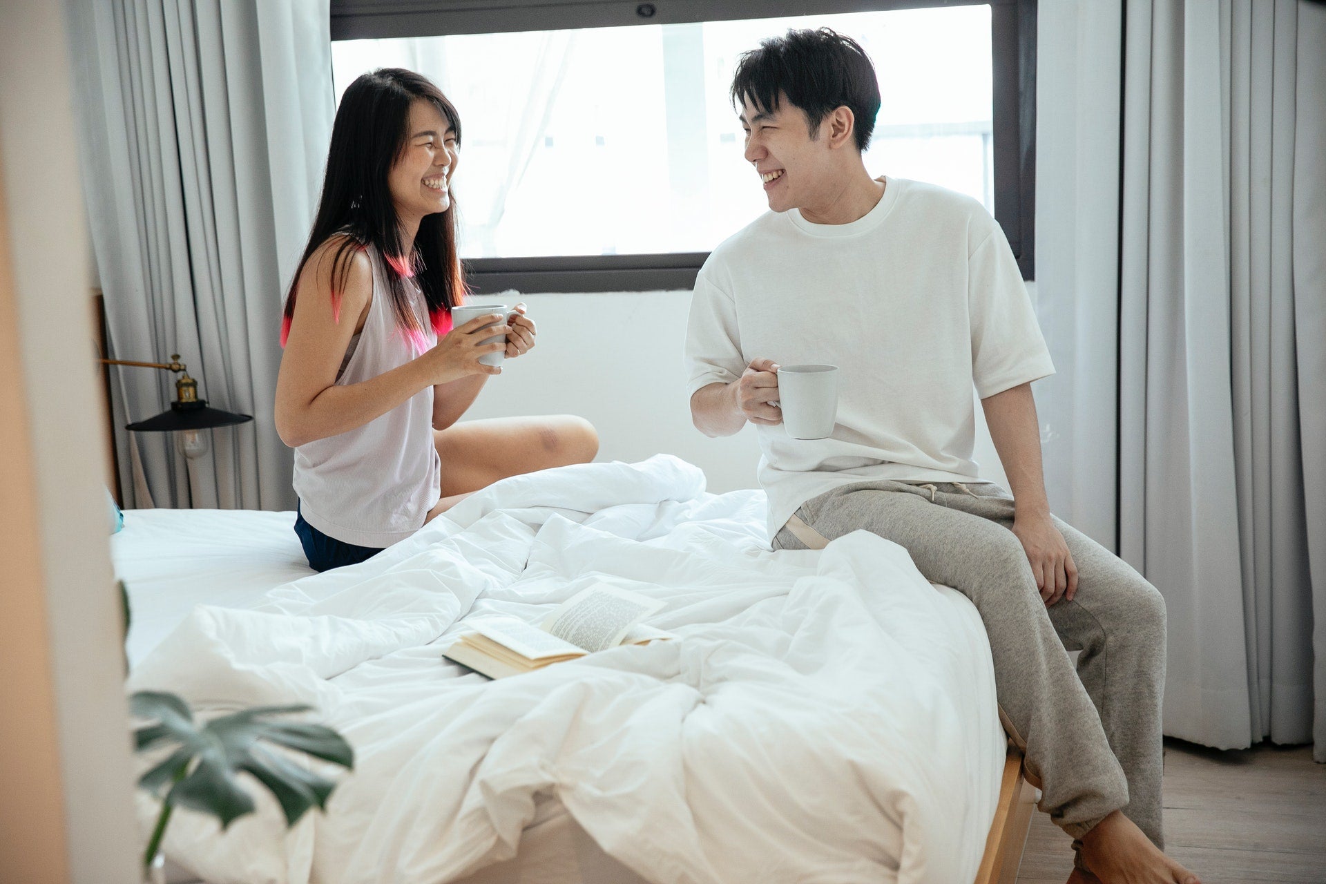 A couple talking to each other and laughing while sitting on a bed in their room, holding mugs
