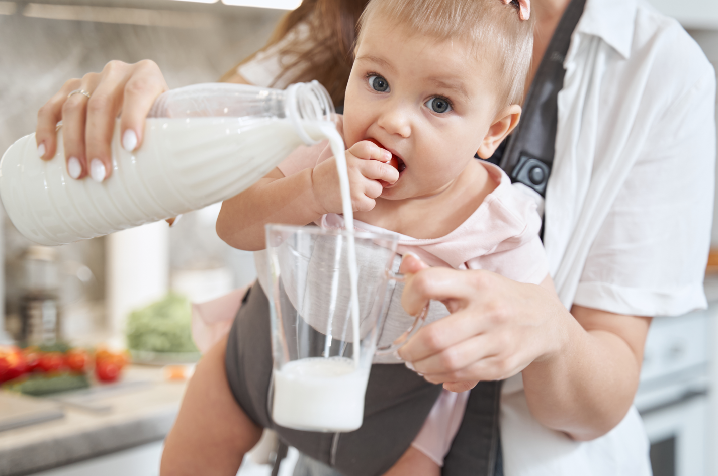 A mother pouring a glass of cow's milk while the baby watches from a baby carrier. Cow's milk is bad for babies.