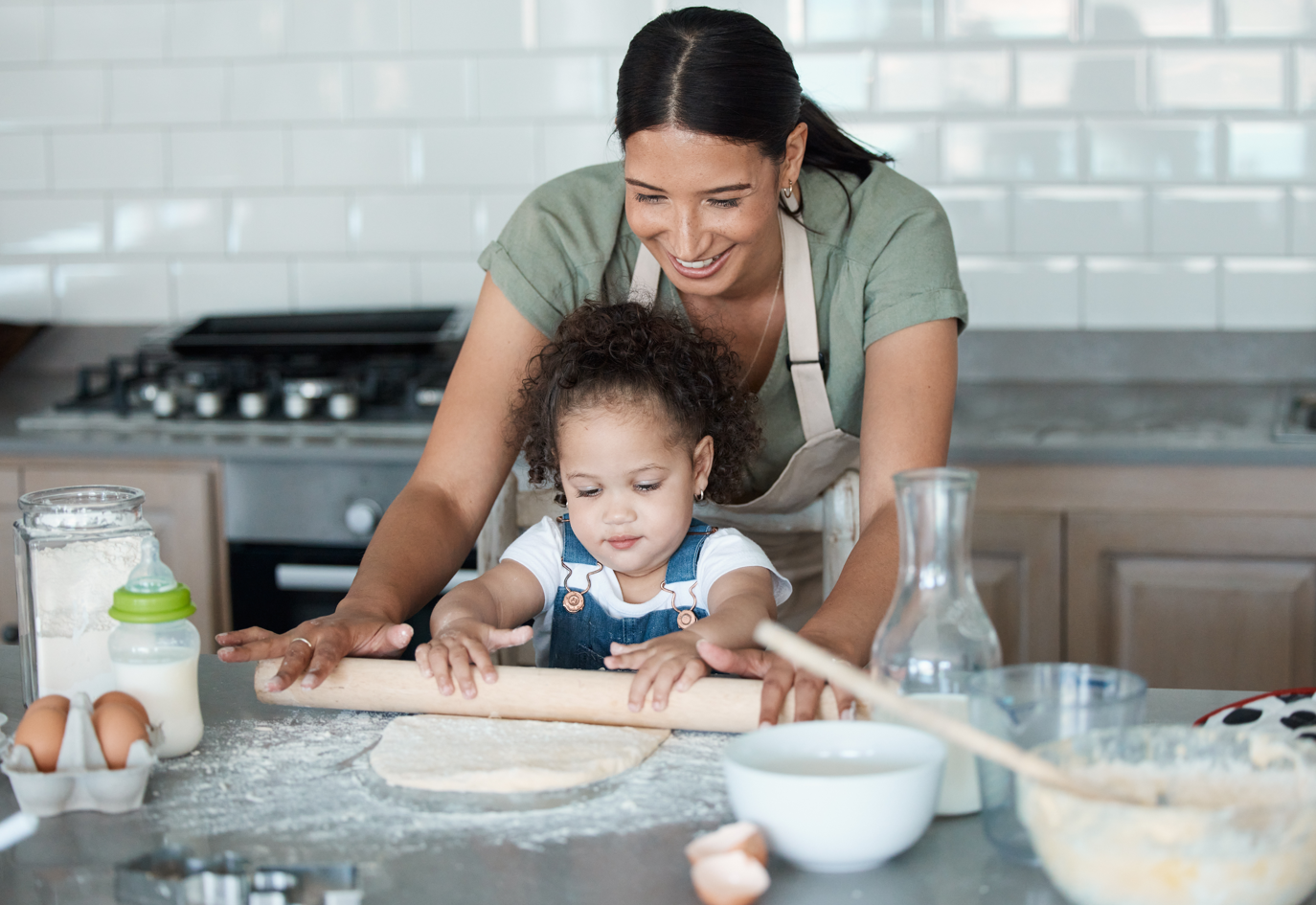 A mother and her toddler stand in the kitchen and roll out dough together. Concept of single parent, baking, toddler, chores.
