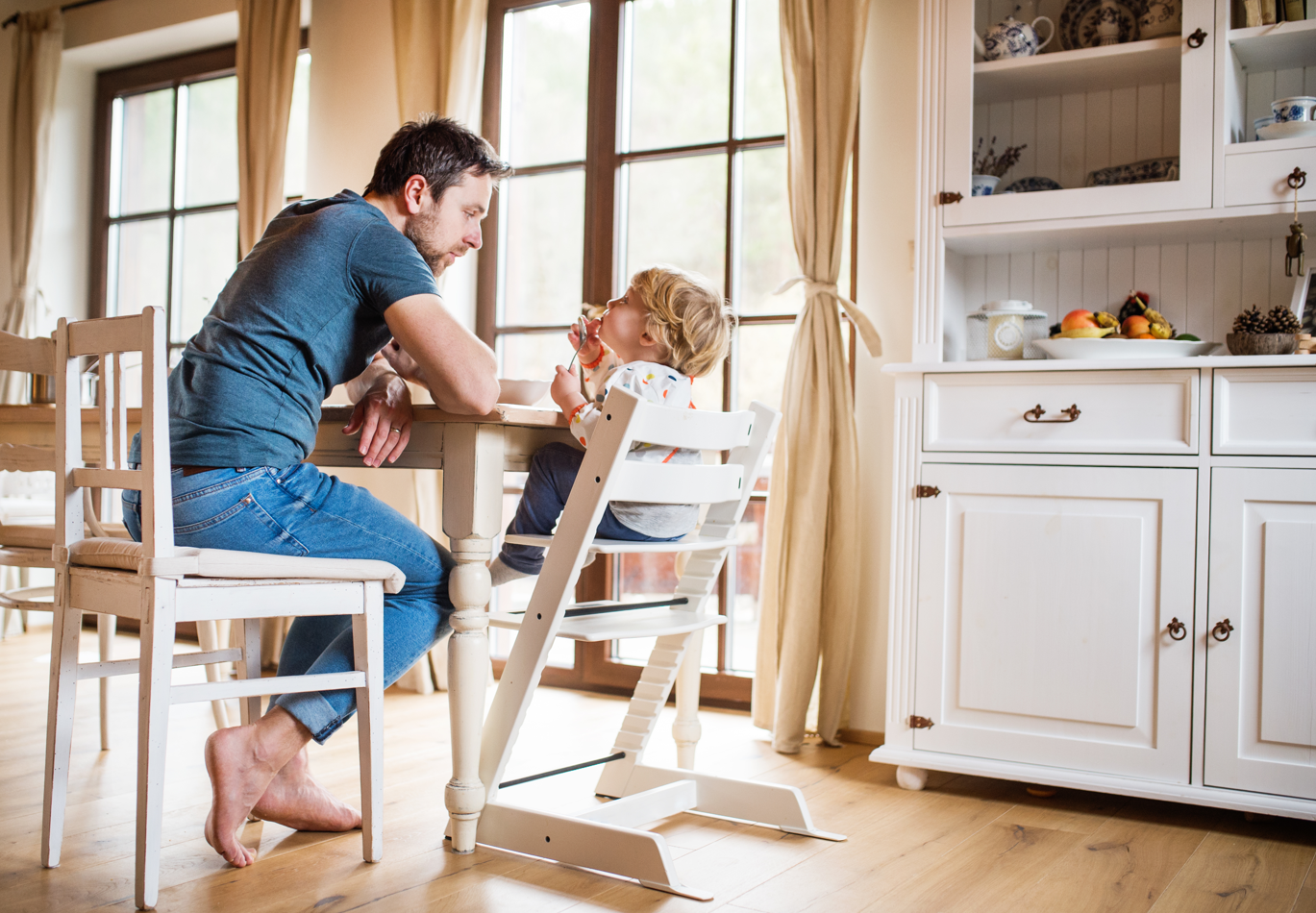 A father and toddler sit at the dining table. Toddler is in a high chair having a meal. Warm, sunny room next to the kitchen. Concept of toddler routines, single parenting, mealtime, toddler tips.