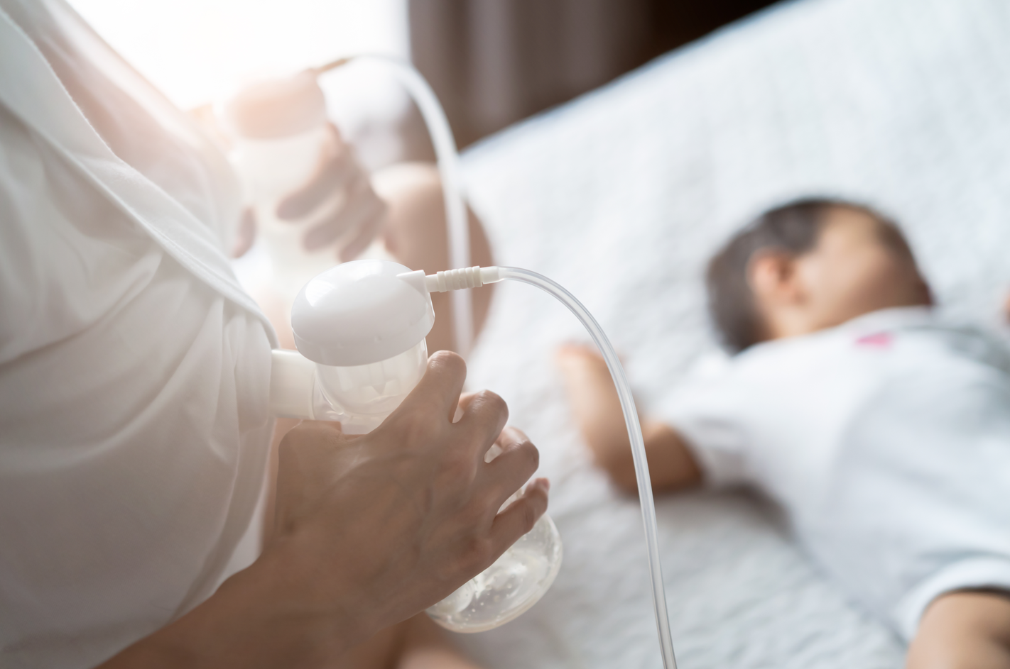 A new mother using breast pumps to relieve her engorged breasts. Face out of view, baby seen sleeping on the bed in the background. Concept of breast engorgement treatments.