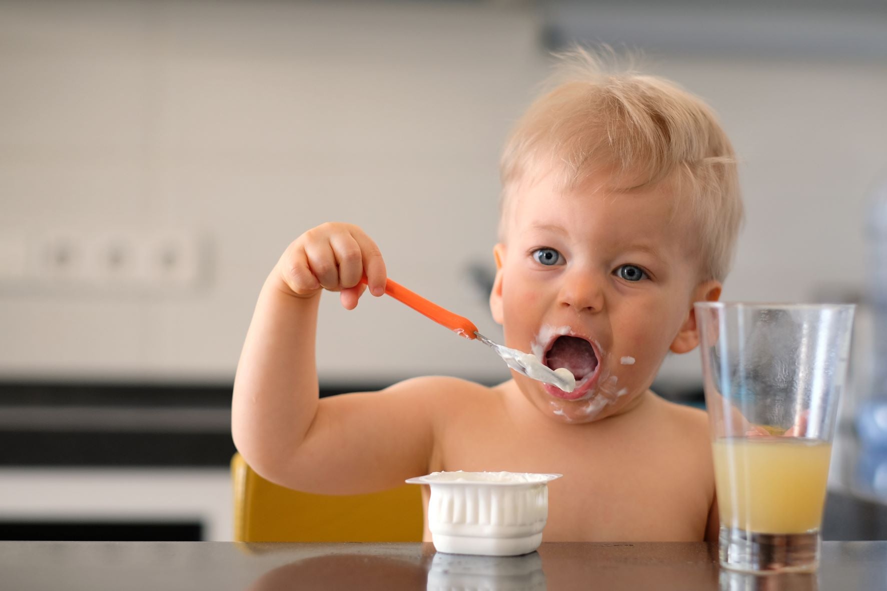 A baby sitting at the kitchen counter and eating yogurt with a spoon. Yogurt is great for babies.
