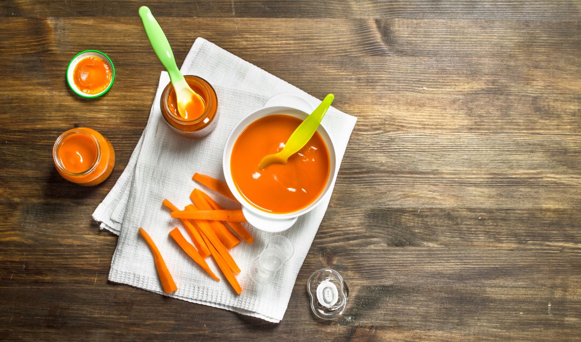 A jar and a bowl of pureed carrot baby food alongside slices of carrots on a wooden surface. Carrots are some of the best foods for babies.