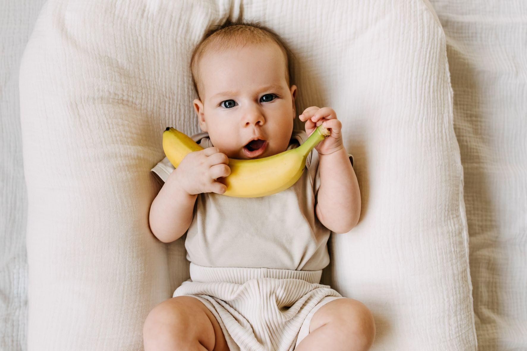 A small baby holds a banana while lying on a bed. Bananas are great first foods for babies.