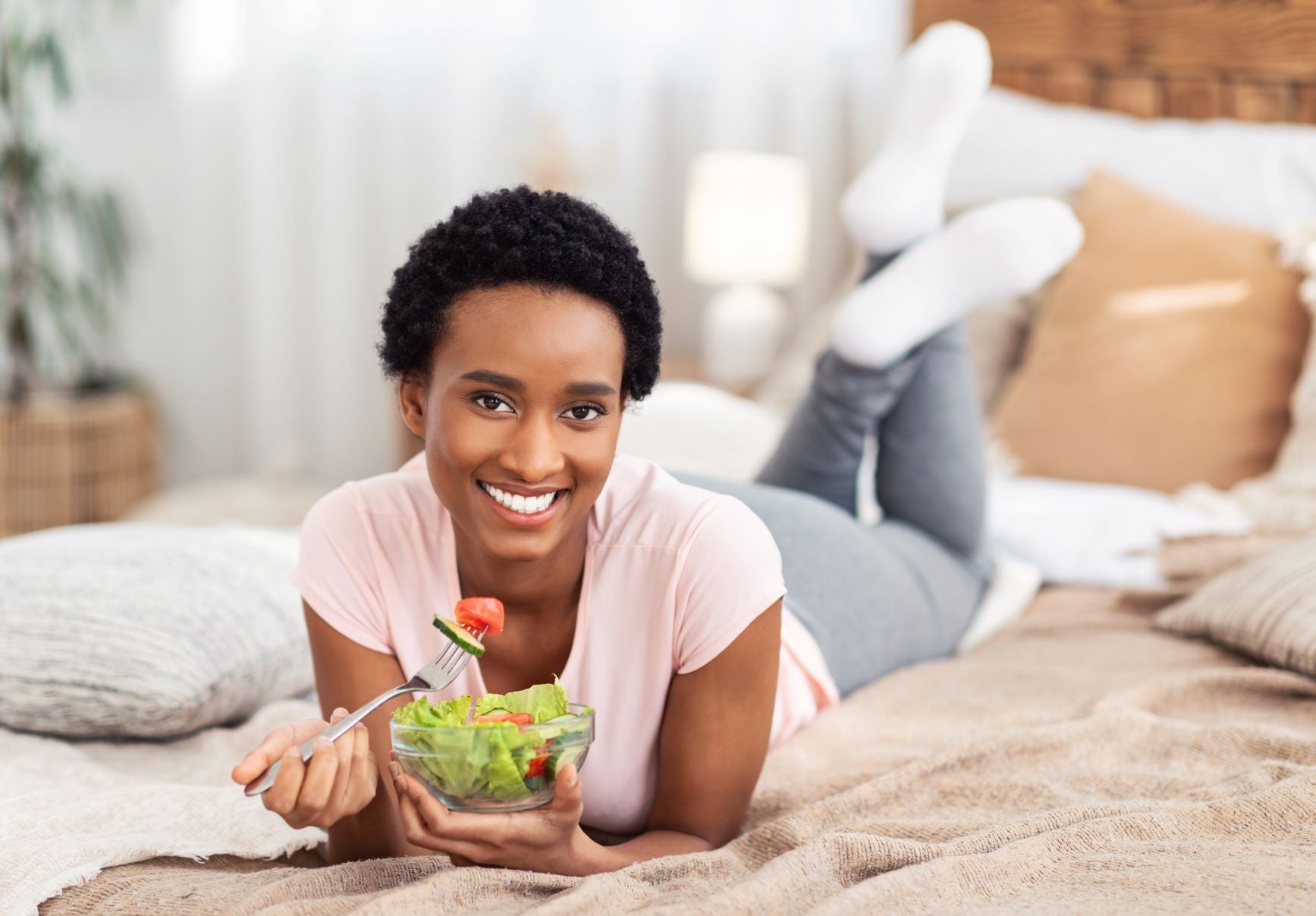 A happy black woman who is a first-time mom lies on the bed and eats a bowl of healthy salad. 