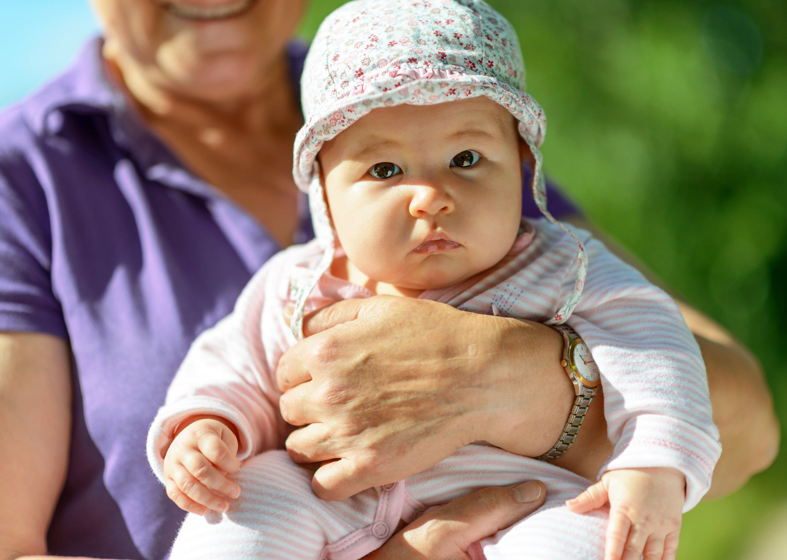A grandmother holds her baby in the sun outside. The baby is fully covered by cotton clothes and a hat so as to be protected from the sun's rays