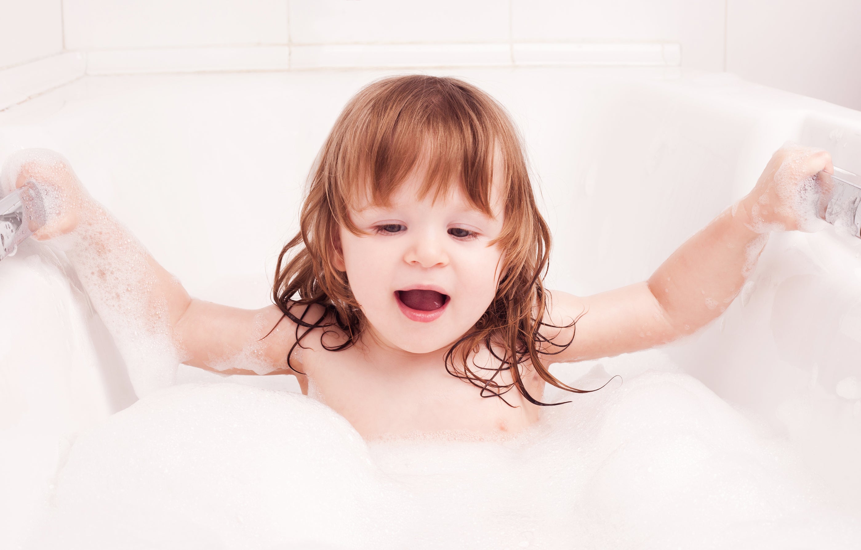 A toddler with long hair taking a bubble bath in a white bathtub.