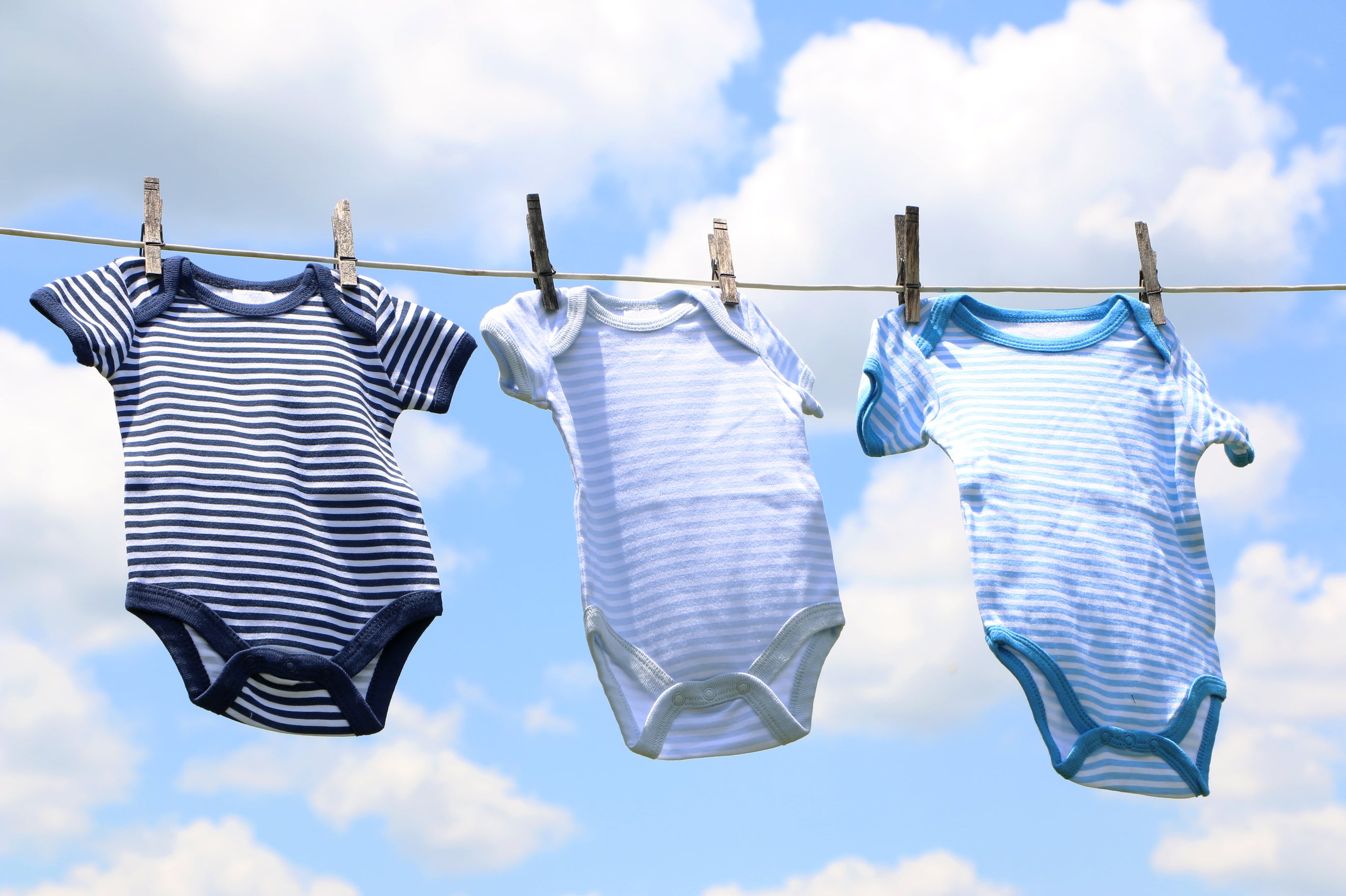 Baby onesies or rompers hanging on a clothing line against a blue sky. If your baby has outgrown their cute clothes, you can turn them into cleaning rags!