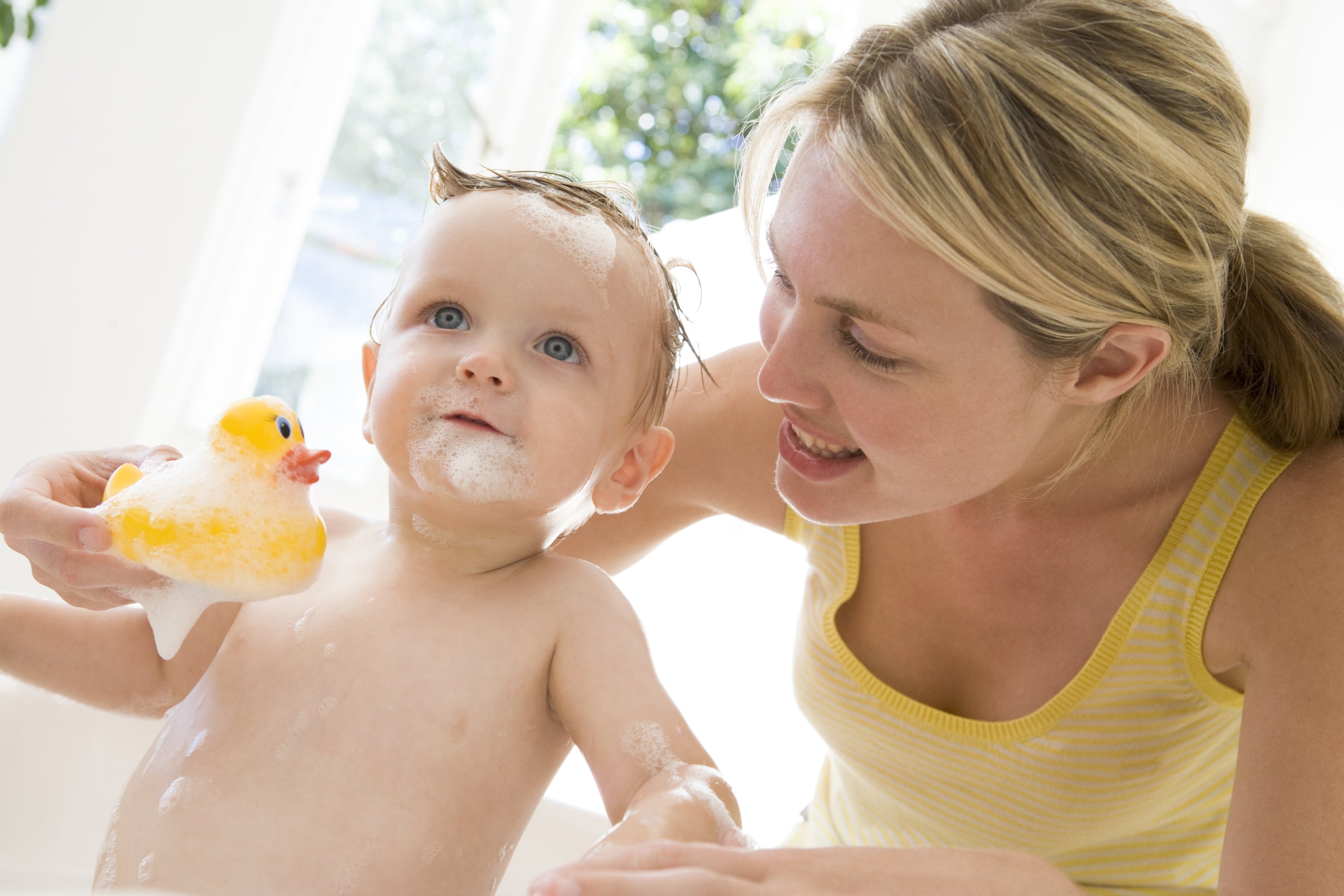 A baby over six months old accompanied by his mother holds a rubber duck and stands in the bubble bath