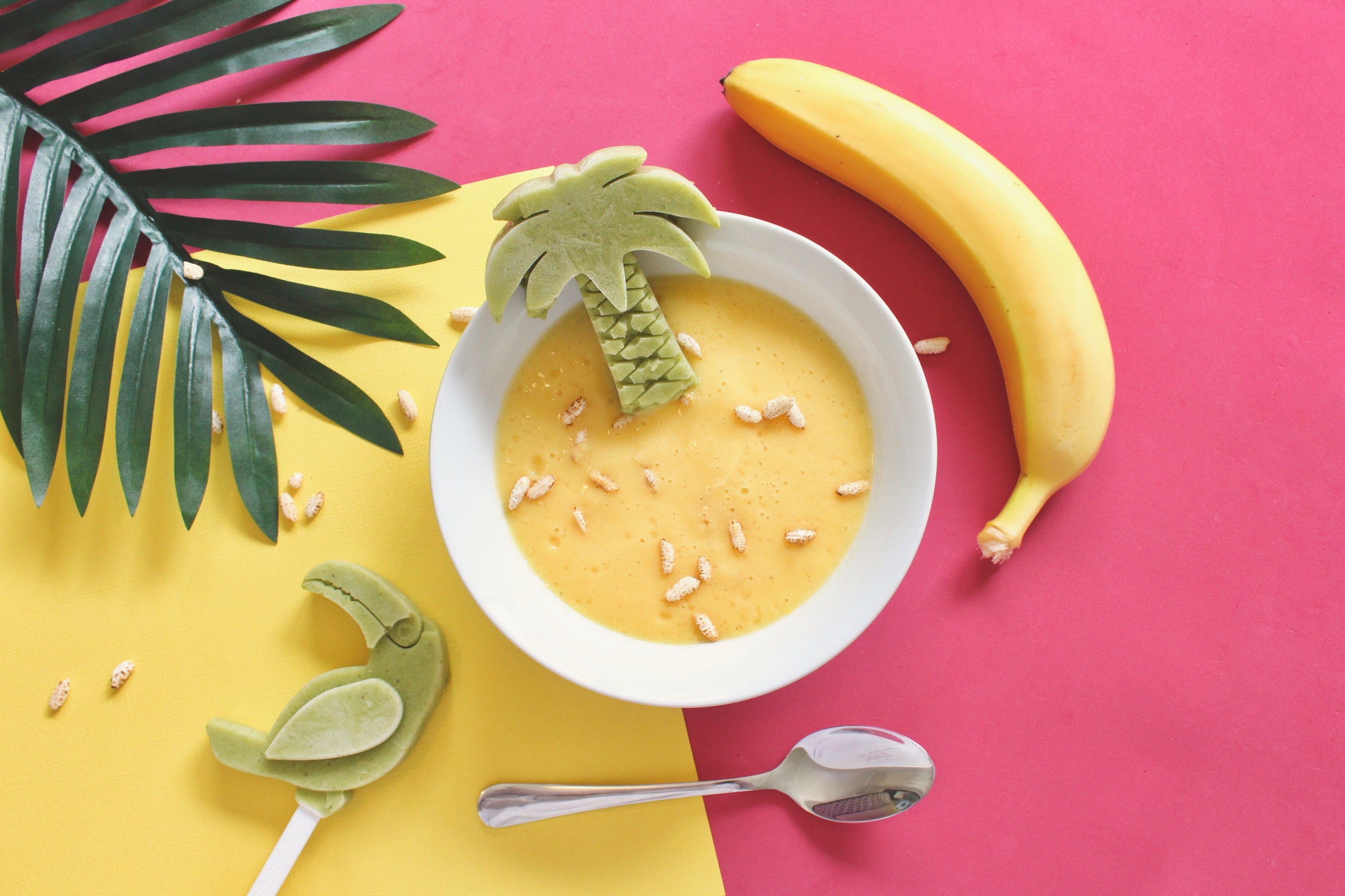 Mashed banana baby food in a bowl against a colorful surface with a banana on the side