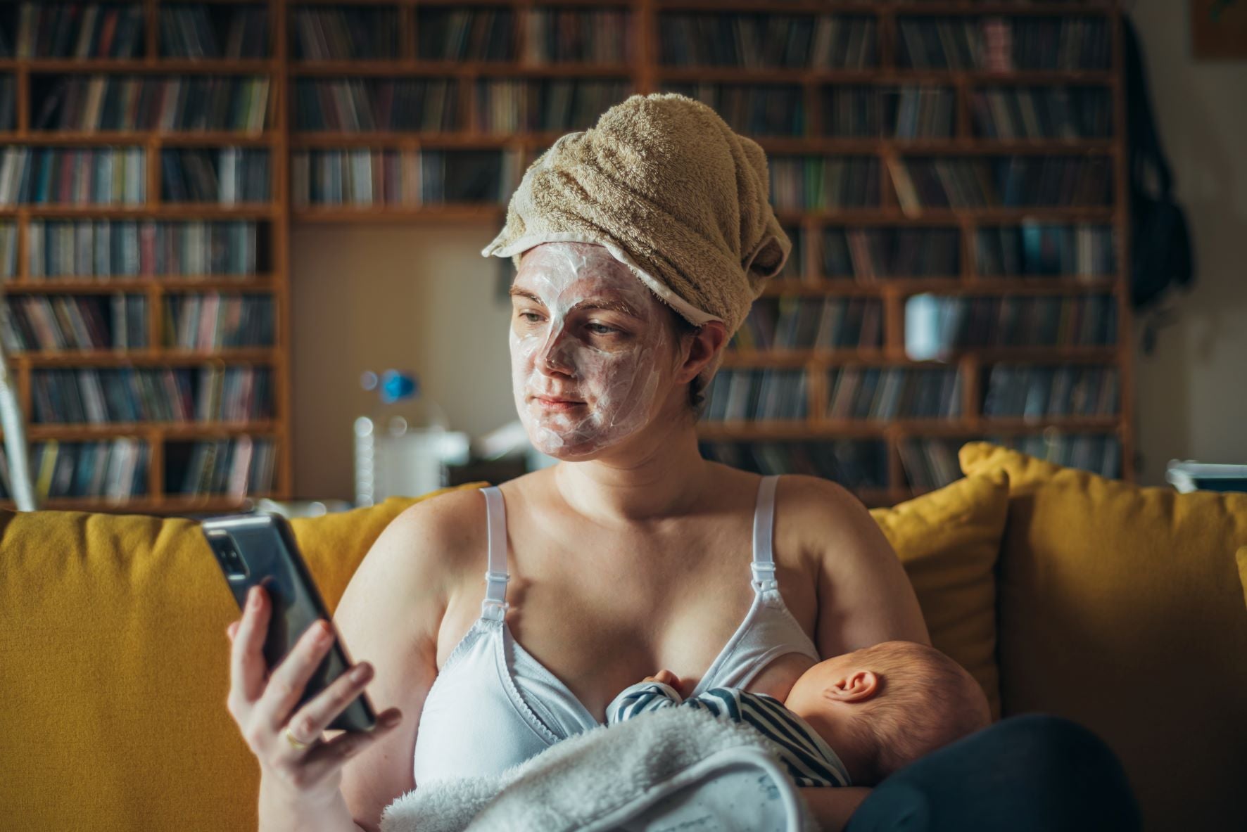 A nursing mother wearing a face and hair mask, scrolling through her phone, and comfortably breastfeeding her newborn baby. 