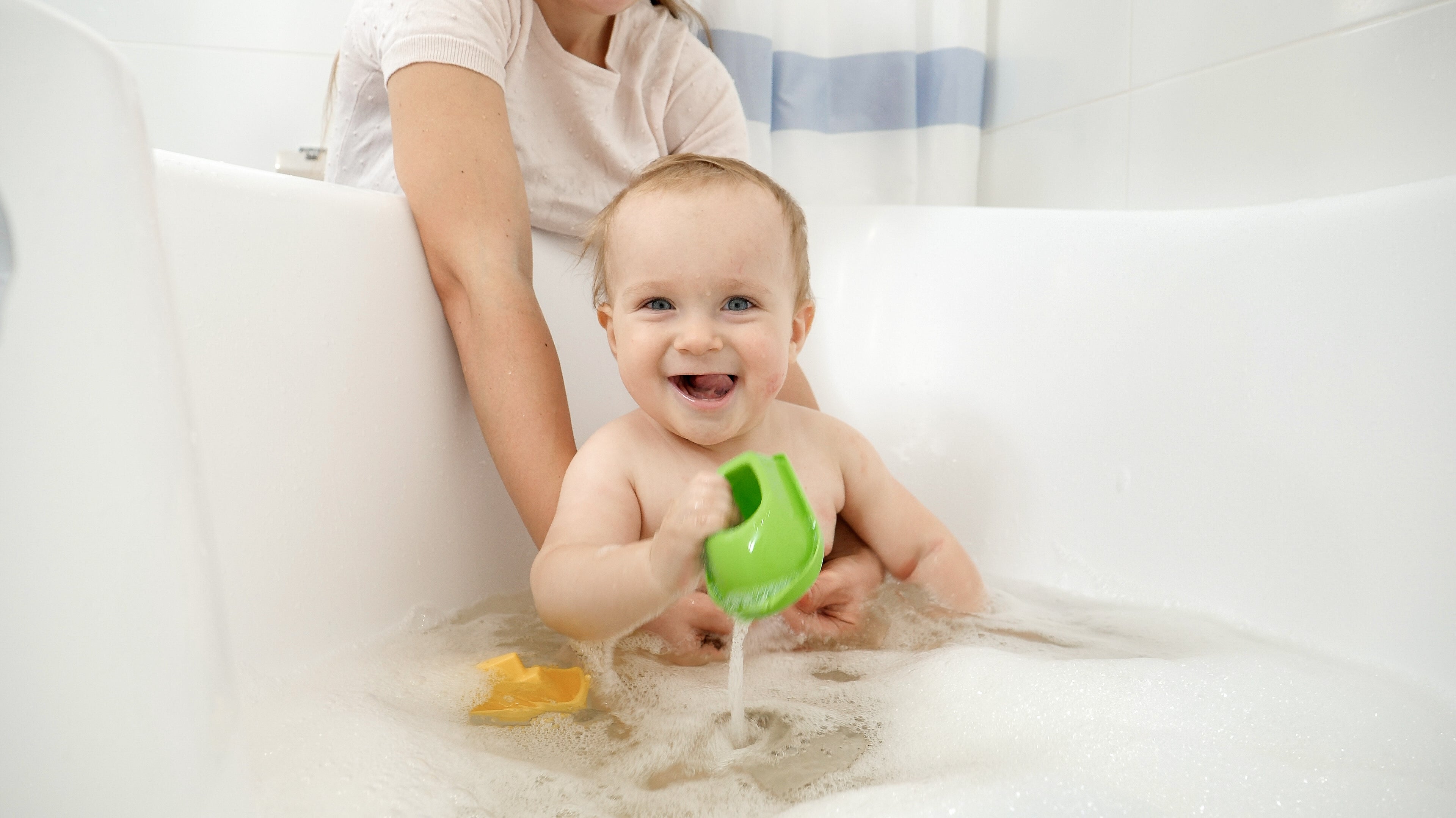 A toddler in a bathtub playing with bath toys and bubbles. Mother's arm is visible in the background.