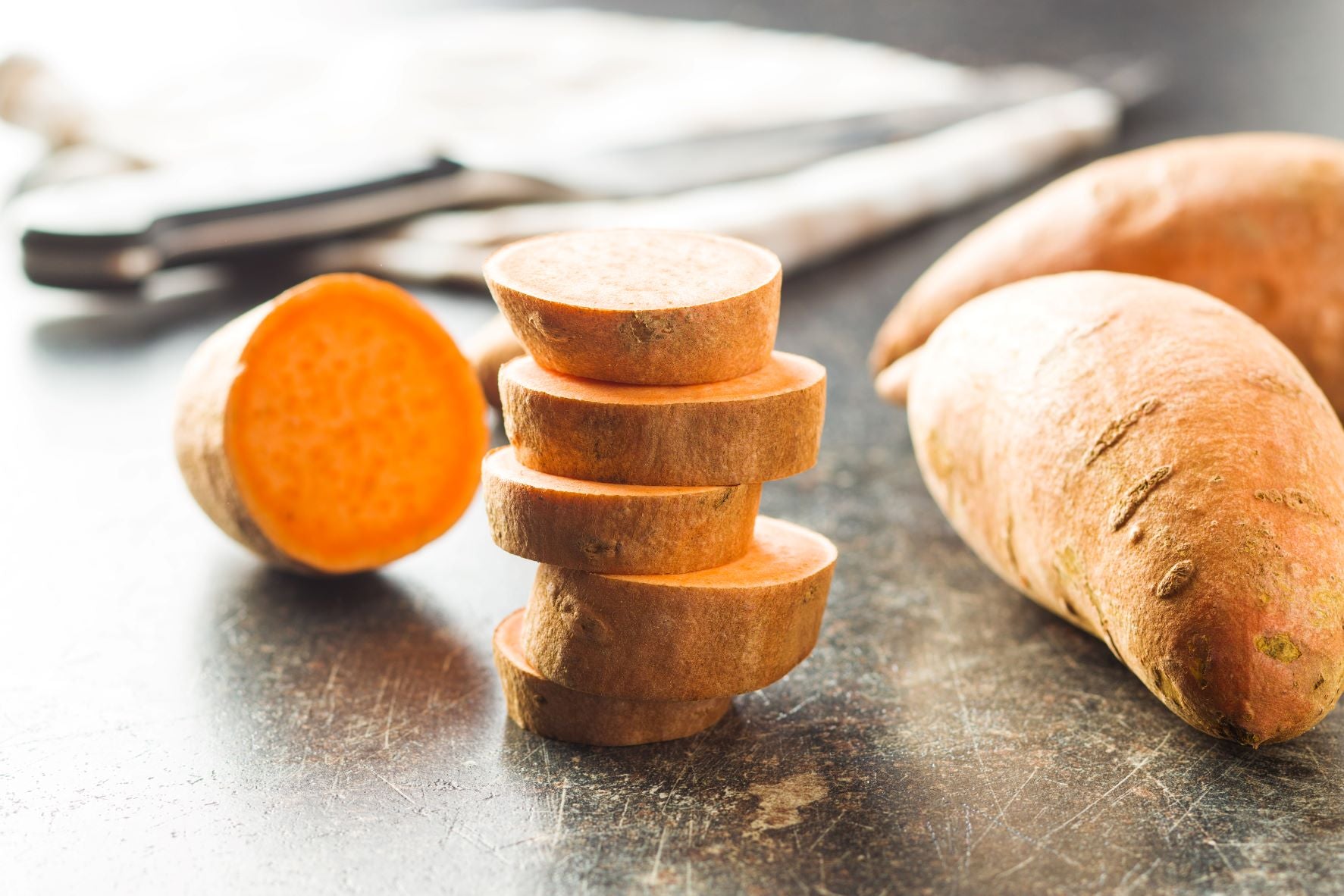 Stacked slices of sweet potatoes alongside a knife and whole sweet potatoes. Babies can have sweet potatoes as they are healthy.