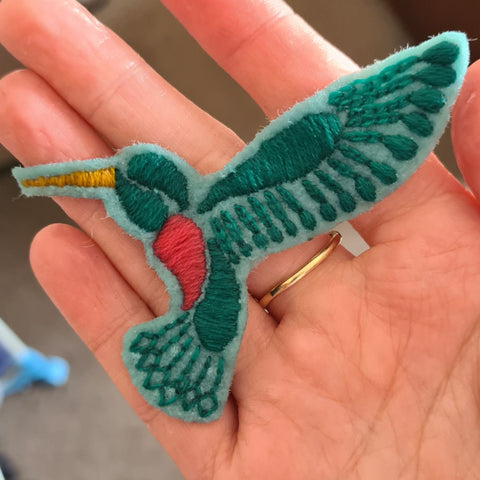 a hand holding an embroidered hummingbird patch, which has just been dipped in water to remove transfer paper