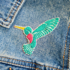 A turquoise hummingbird design has been hand stitched onto a piece of felt, then stitched onto a denim jacket