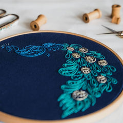 An embroidery hoop containing blue fabric stitched with a blue, teal, and gold peacock design.